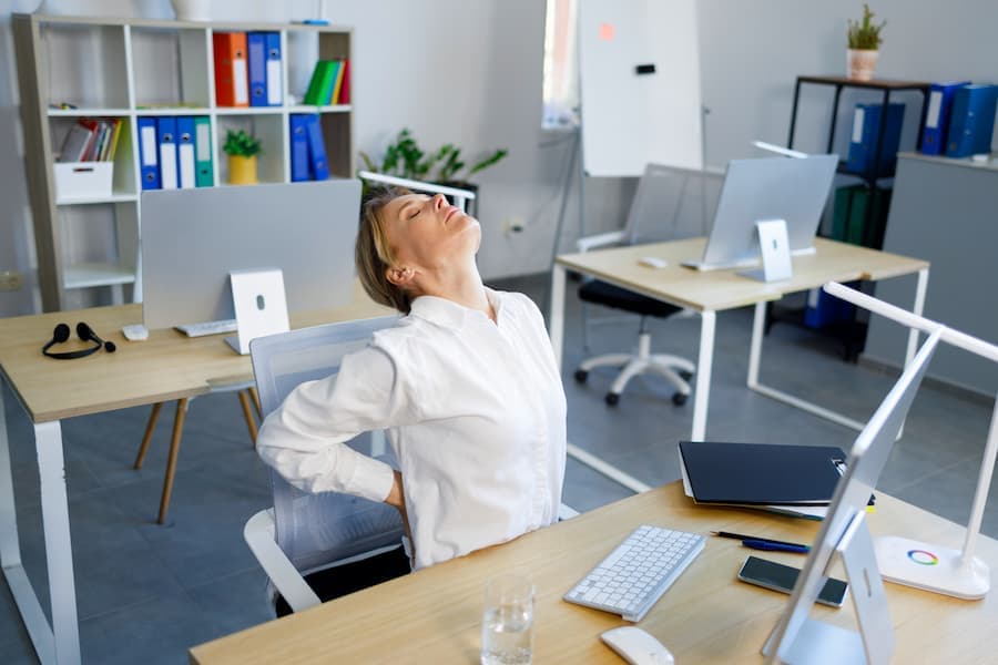 mulher sentada na mesa de trabalho com as mão na lombar sentindo dor
