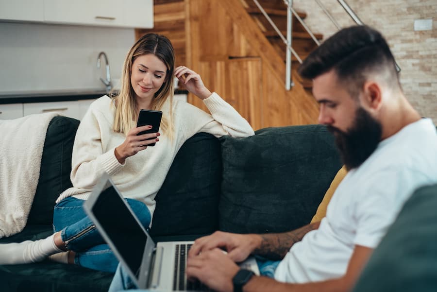 Casal de jovens sentados no sofá, ela mexendo no celular, ele no note book