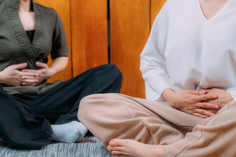 imagem de duas mulheres, do pescoço para baixo, sentadas, em posição de ioga, com as mãos na barriga, treinando a respiração