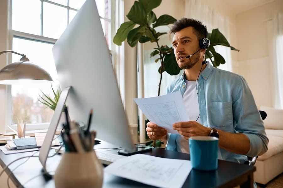 Homem trabalha em frente ao computador usando fone de ouvido, na sala de casa