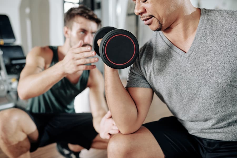 Homem levanta peso auxiliado por personal trainer