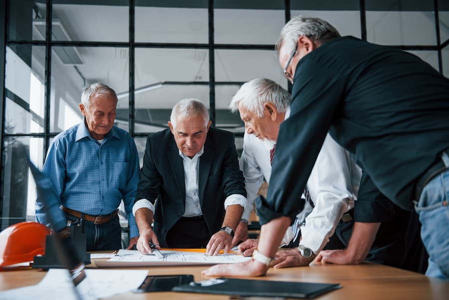 Grupo de homens maduros, todos de cabelos brancos, discutem projeto em ambiente de trabalho