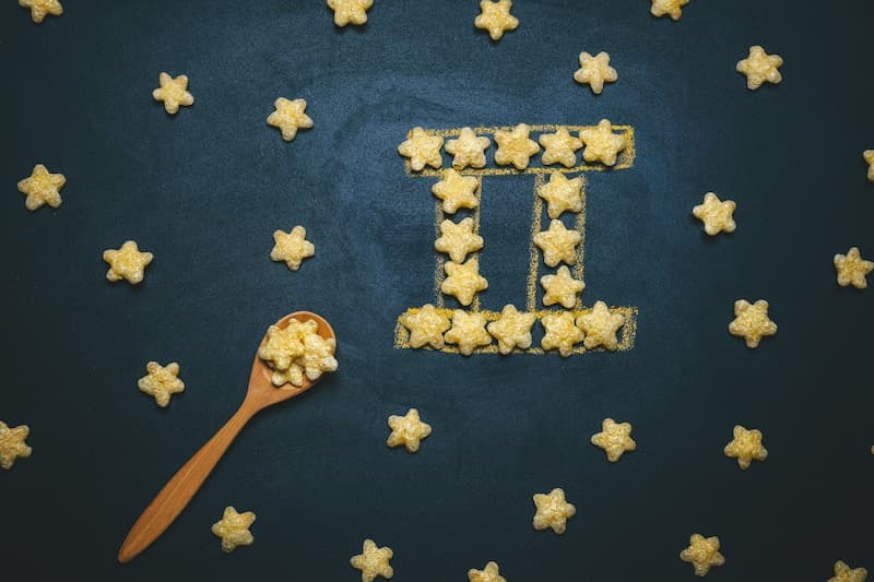 Estrelas de cereal dispostas sobre uma superfície escura formam o símbolo do signo de Gêmeos. Algumas estrelas estão espalhadas ao redor e uma colher de madeira contendo mais estrelas está posicionada no canto inferior esquerdo da imagem.