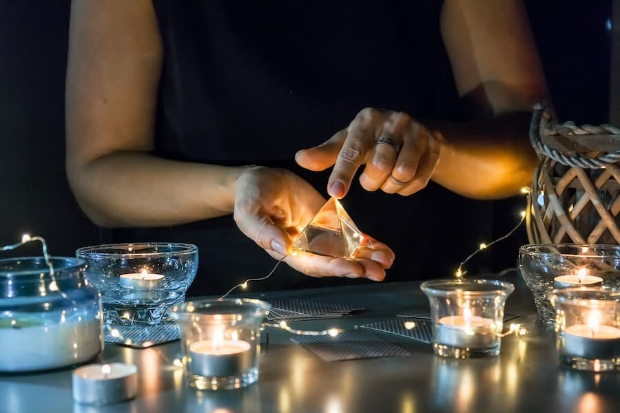 mãos de mulher manipulam oirâmide de cristal em meio a velas e cartas
