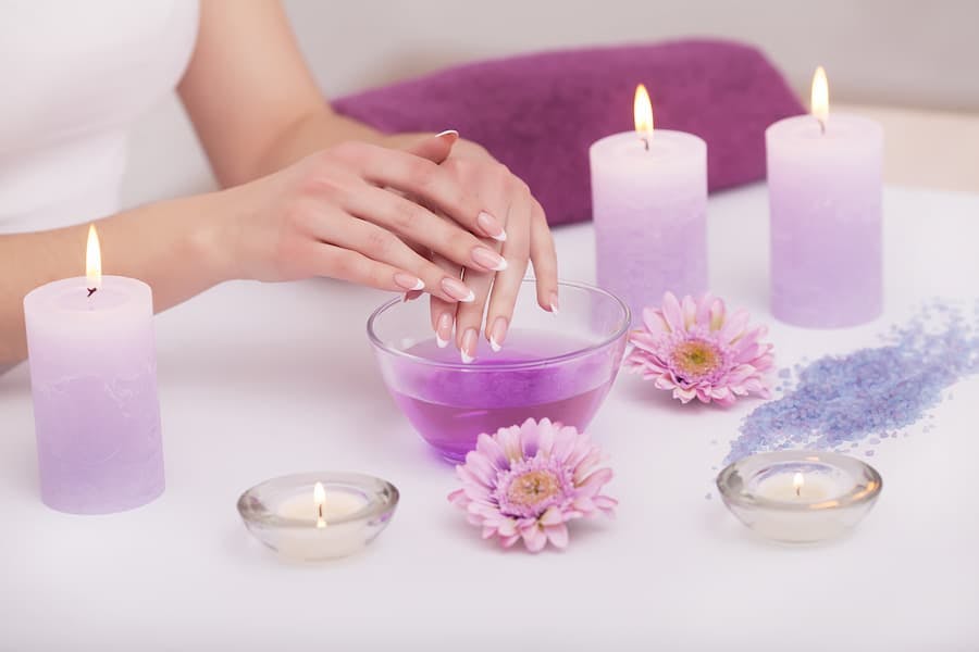 Mãos de mulher mergulando numa tigela de água, ao lado de velas e flores em tons de branco e lilás