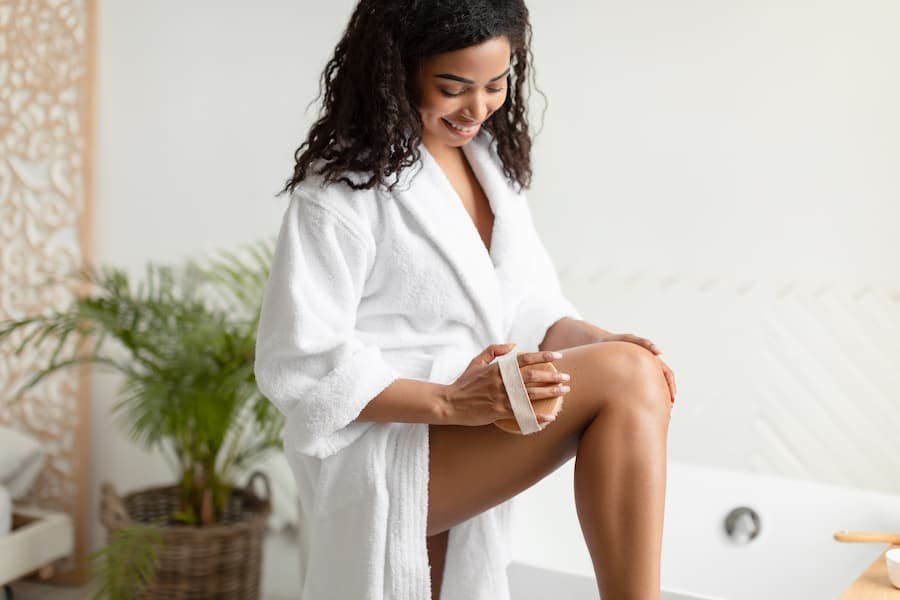 Mulher jovem com roupão de banho faz massagem na coxa com uma escova