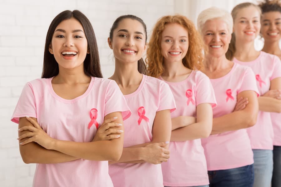 Mulheres enfileiradas com camiseta e fita rosa do Outubro Rosa em atenção ao câncer de mama