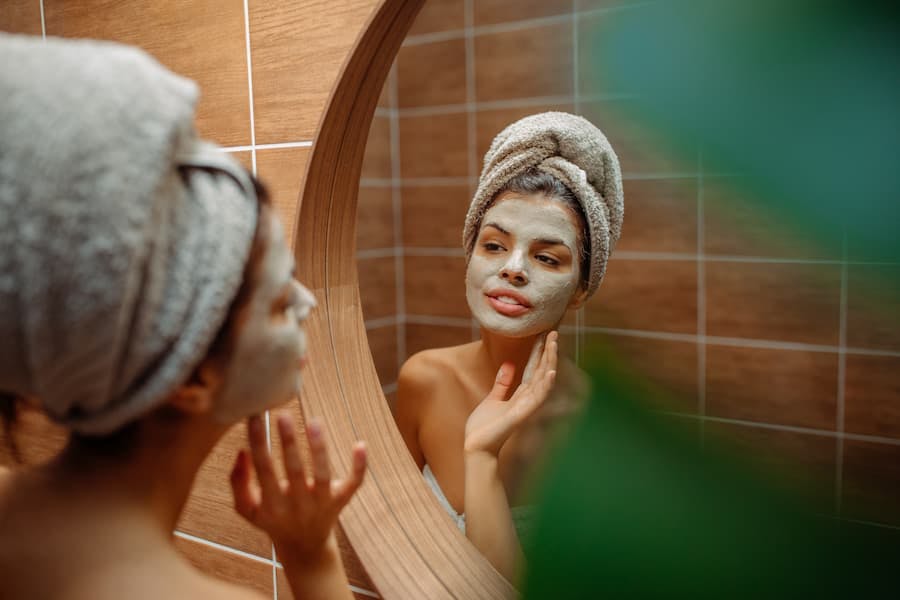 Mulher com toalha na cabeça, em frente ao espelho, passa creme no rosto