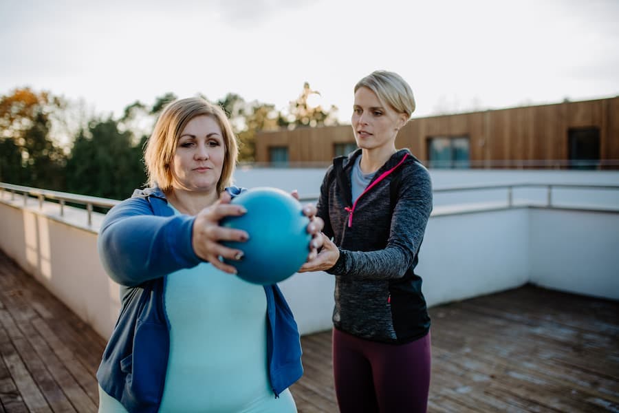 Mulher acima do peso segura bola de exercícios sob supervisão de uma profissional